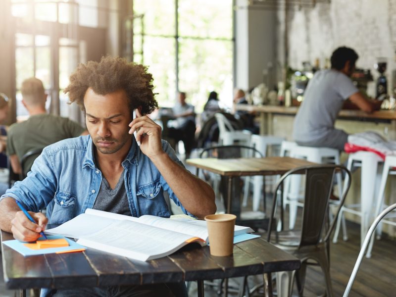 Mindful dark-skinned male student wearing casual clothing preparing for exams sitting at cafe table before lessons, reading information in textbook and talking on phone with his coach asking questions